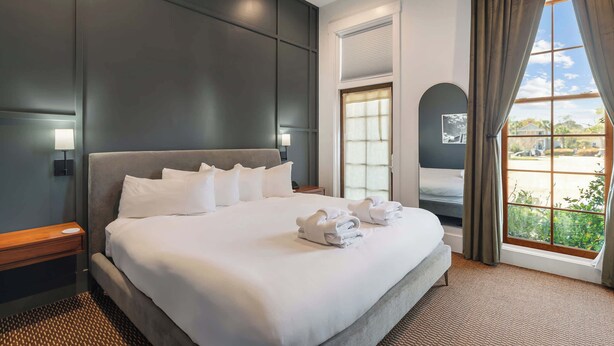 Ein Hotelzimmer mit einem großen Bett, einem Nachttisch, einem Fenster mit Vorhängen und Blick ins Freie.