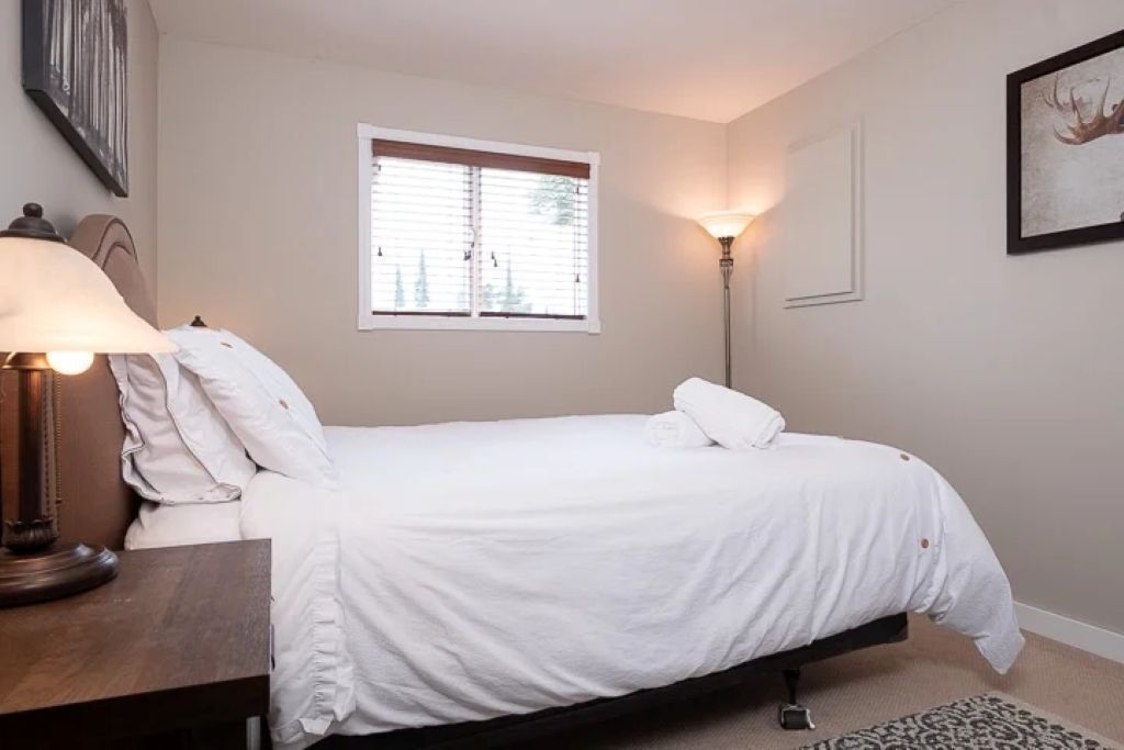 A bedroom with a bed, a nightstand, a lamp, a window with blinds, and a framed picture on the wall.