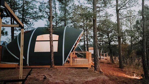 En campingplats i ett skogsområde med ett stort, svart tält med ett vitt fönster och en träveranda.