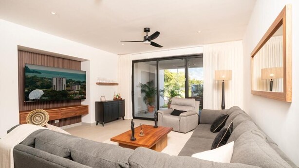 A modern living room with a grey sofa, a wooden coffee table, a television mounted on the wall, a ceiling fan, and a sliding glass door leading to an outdoor area. - Stunning Condo at The Pearl in Jaco Beach 6B (Jacó)