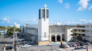 Una chiesa con un'alta torre e una croce, circondata da un parcheggio e altri edifici.