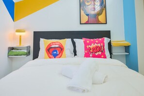 Een bed met witte beddengoed, een kussen met een rood-geel ontwerp, een kussen met een blauw-wit ontwerp, en een kussen met een roze-geel ontwerp.