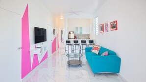A modern living room with a blue sofa, a TV, and a kitchen in the background. - Cozy Escape | Steps from Beach (San Juan)