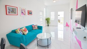 A modern living room with a teal sofa, a glass coffee table, and a flat-screen TV. - Cozy Escape | Steps from Beach (San Juan)