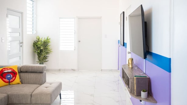 A modern living room with a grey sofa, a TV mounted on the wall, and a potted plant. - Modern Retreat By The Beach (San Juan)