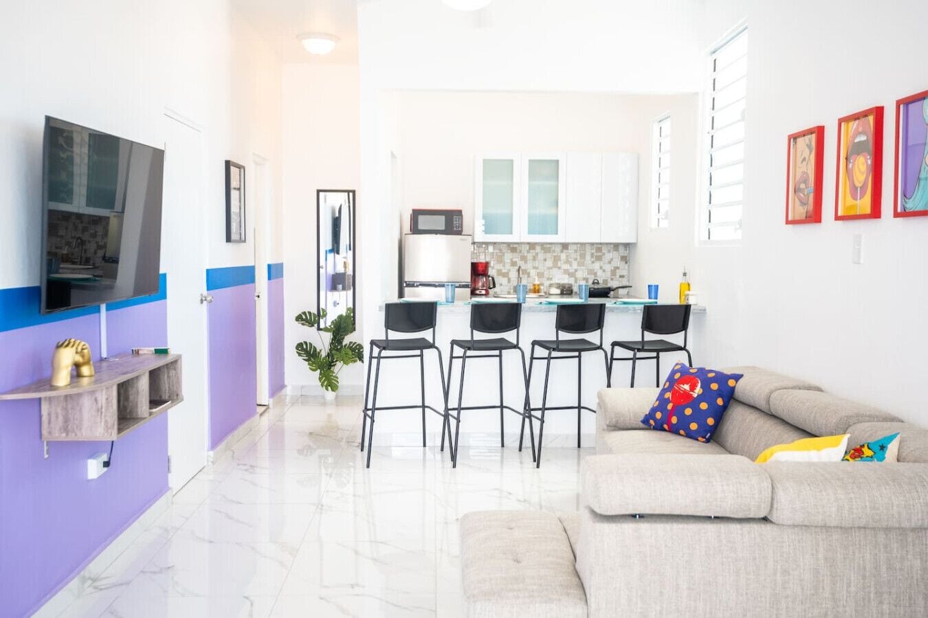A modern living room with a sofa, a TV, a kitchen area with bar stools, and a dining table.