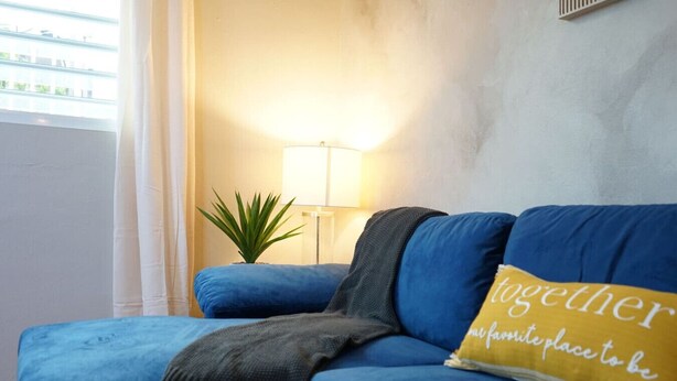 Ein blaues Sofa mit einem gelben Kissen, einer kleinen Pflanze und einer Stehlampe in einem Zimmer mit Fenster und schlichter Wand.