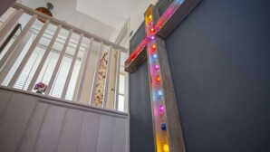 Ein hölzernes Kreuz mit bunten Lichtern, ein weißes Geländer und ein Fenster mit blumigen Vorhängen.