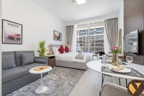 Una habitación de hotel moderna con un sofá gris, una cama con almohadas rojas, una mesita redonda pequeña y un comedor con una mesa y sillas blancas.