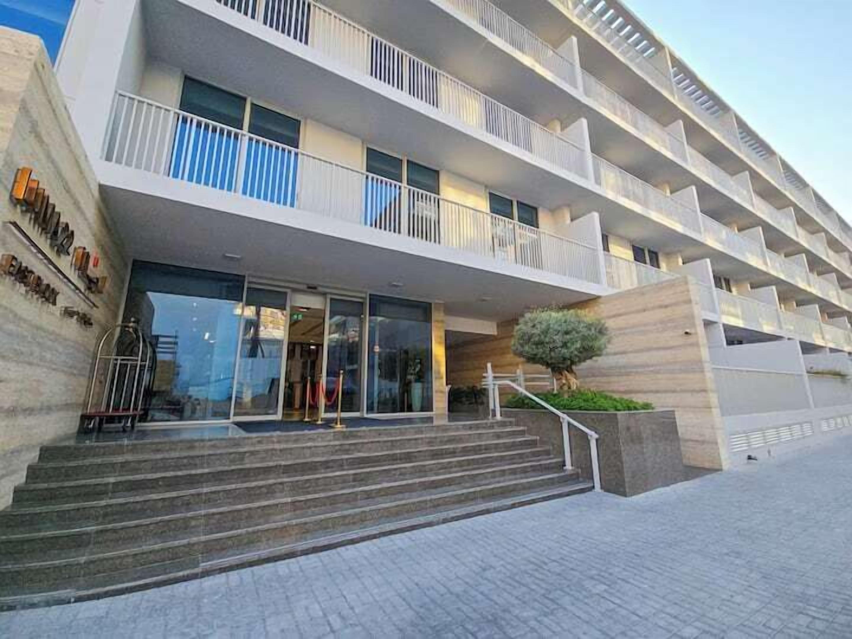 A multi-story hotel with a modern design, featuring a central staircase leading to the entrance, large windows, and a small tree in the foreground.