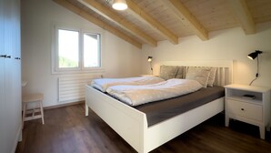Une chambre avec un lit blanc, une table de chevet, un petit tabouret, une fenêtre et des poutres en bois au plafond.