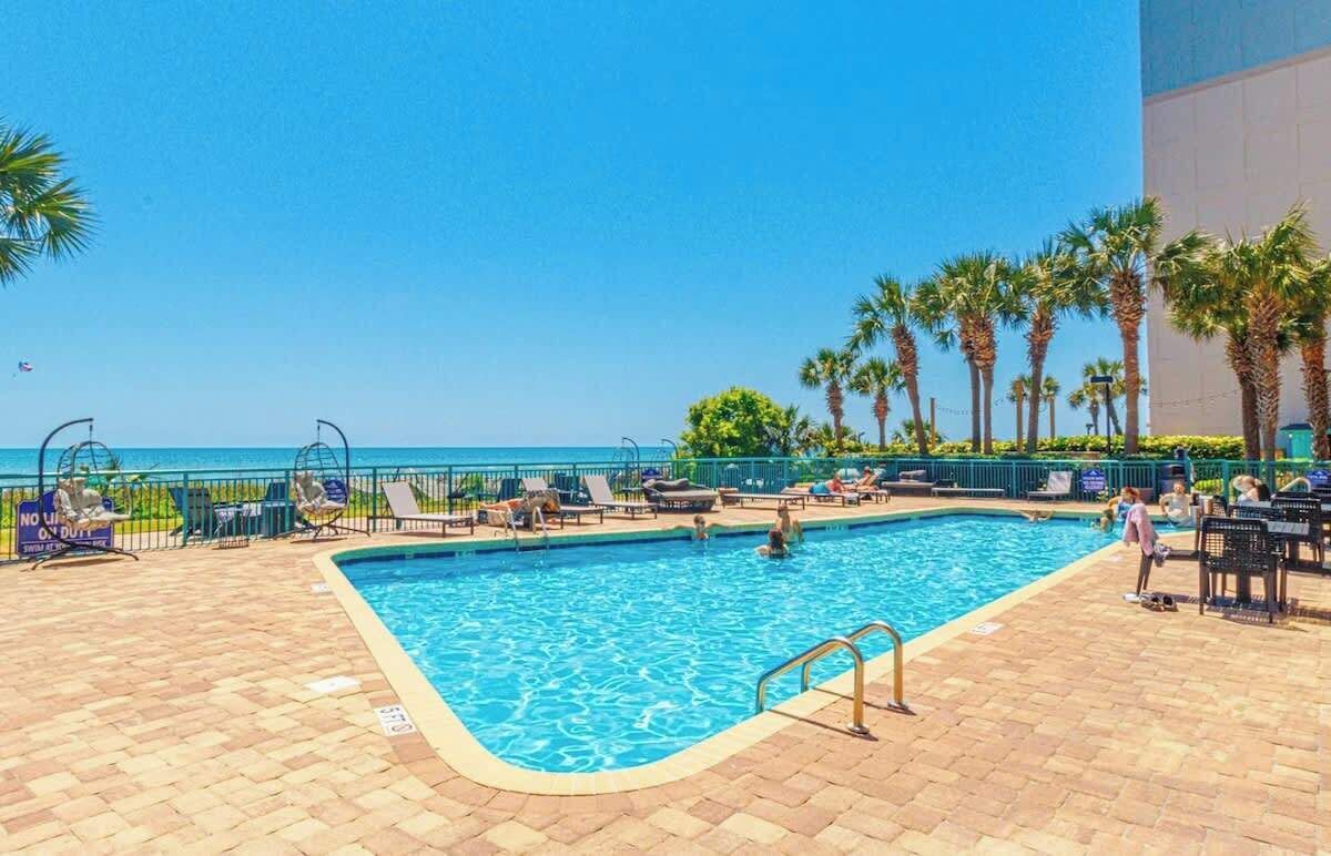 A pool area with lounge chairs and palm trees, overlooking a clear blue sky and ocean.
