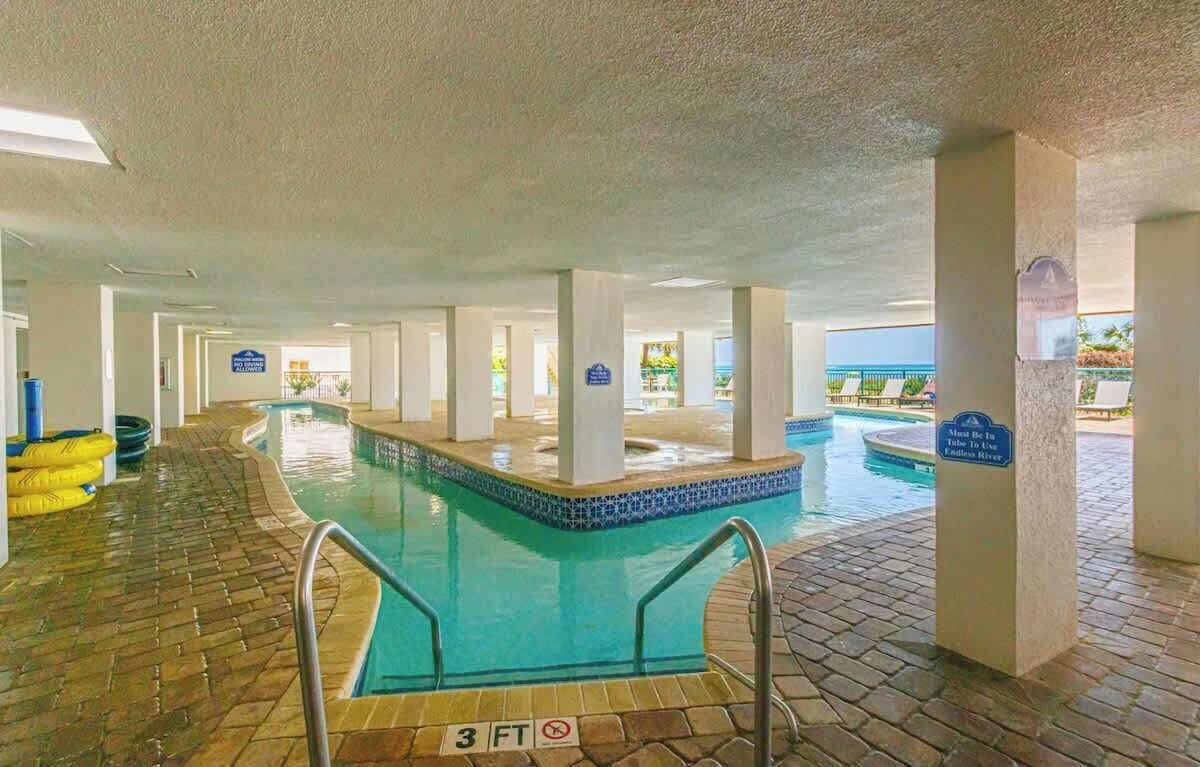 Área interna com piscina, sinalização clara, chão em mosaico e vista para o oceano.