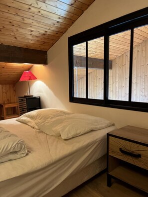 Ein Schlafzimmer mit Holzbalkendecke, einem Bett mit weißer Bettwäsche, einer roten Lampe und einem Fenster mit Holzläden.