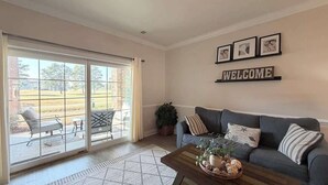 Un salon comprenant un canapé, une table basse agrémentée d’objets décoratifs, une porte-fenêtre coulissante donnant sur un espace extérieur aménagé, et une pancarte « BIENVENUE » accrochée au mur.