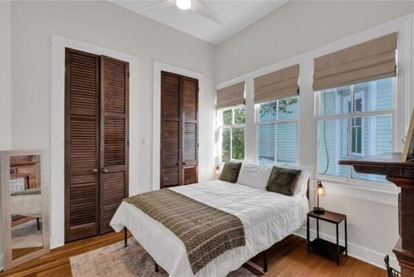 A bedroom with a large bed, two windows with blinds, a nightstand, and a wooden floor.