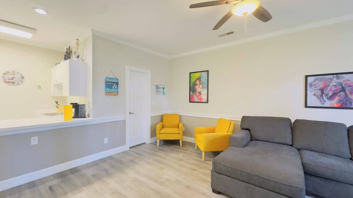 A living room with a gray sofa, two yellow armchairs, a ceiling fan, and a white cabinet with kitchen items.
