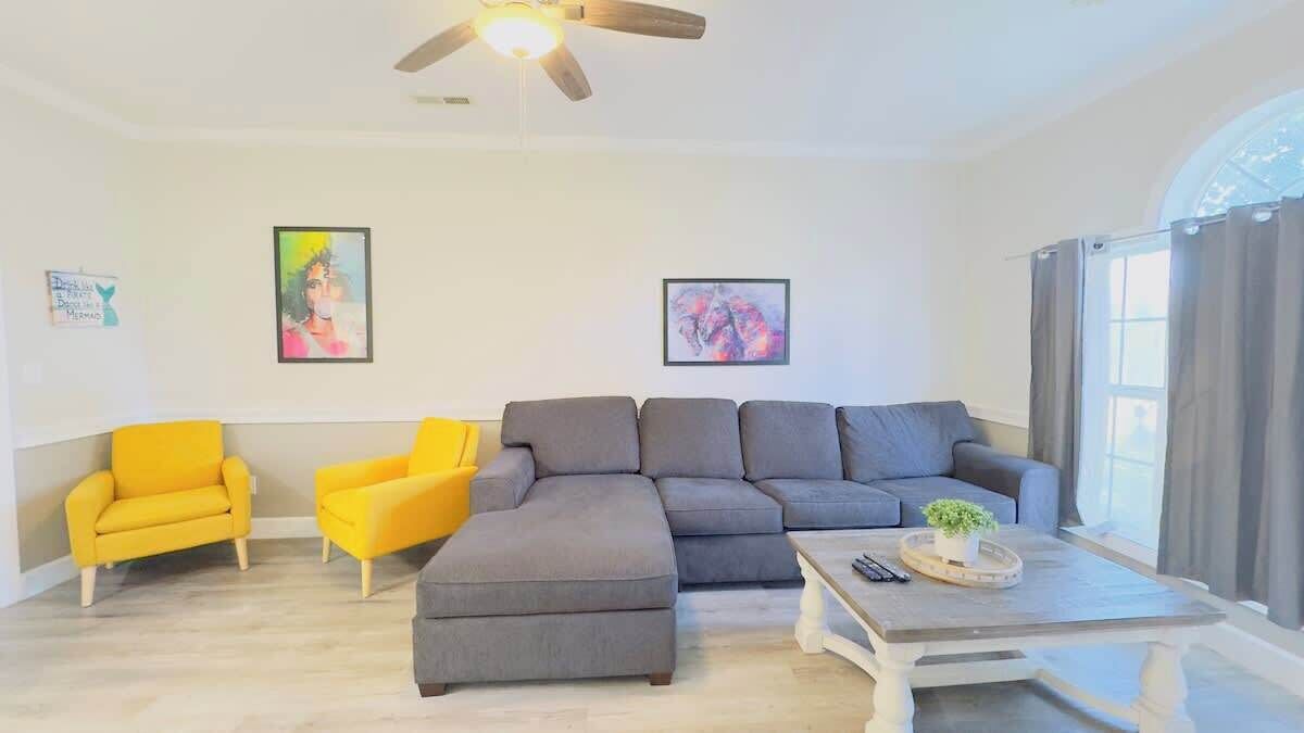 Ein Wohnzimmer mit einem grauen Ecksofa, zwei gelben Sessel, einem Couchtisch mit Pflanze und Kunstwerken an den Wänden.