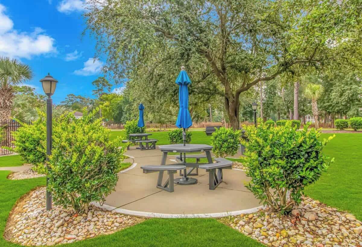 Un parco con tavoli da picnic e ombrelloni blu, circondato da alberi e un prato curato.