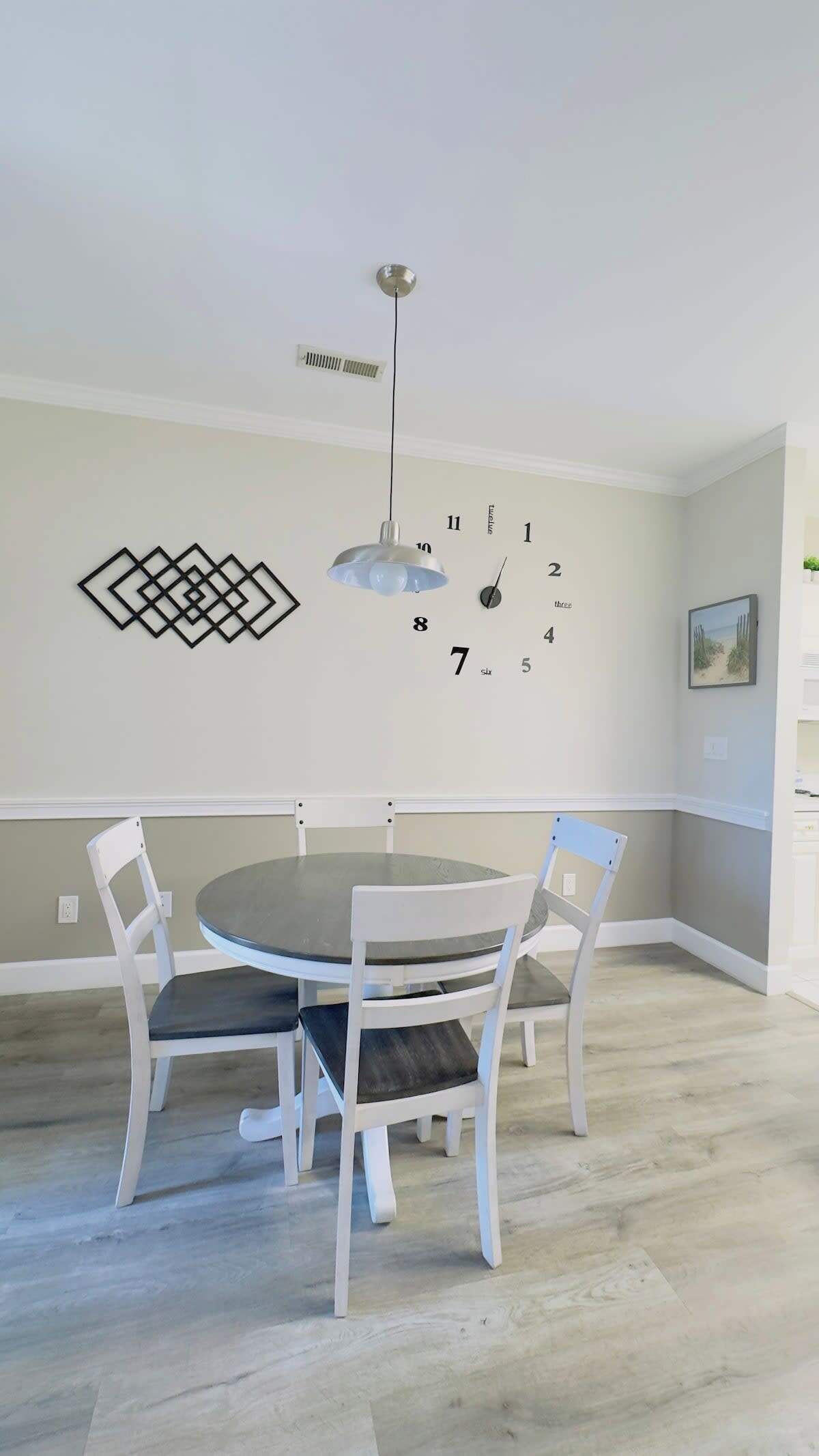 Een eethoek met een ronde tafel, vier witte stoelen, een hanglamp, een wandklok en een geometrisch kunstwerk.