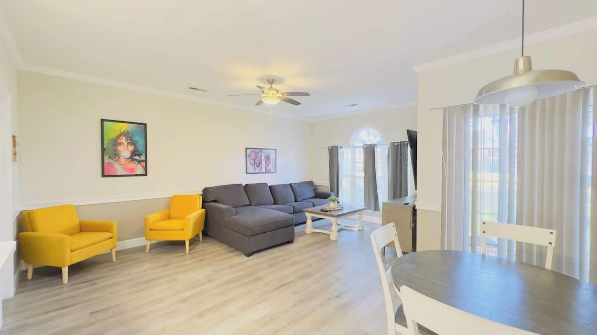 A modern living room with a grey sectional sofa, two yellow armchairs, a dining table with chairs, and a ceiling fan.