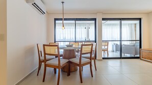 A dining area with a round table and chairs, a hanging light fixture, and a large window with curtains. - APTO Próximo à Praia | 4 Suítes | Varanda Gourmet | San Martin | Destino BSM 303C (Barra de São Miguel)
