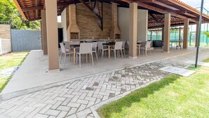 An outdoor patio with a brick barbecue area, tables, and chairs. - APTO Próximo à Praia | 4 Suítes | Varanda Gourmet | San Martin | Destino BSM 303C (Barra de São Miguel)