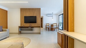 A modern living room with a flat-screen TV, wooden panel wall, a dining area with chairs, and a large window. - APTO Próximo à Praia | 4 Suítes | Varanda Gourmet | San Martin | Destino BSM 303C (Barra de São Miguel)