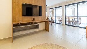 A modern living room with a flat-screen TV mounted on a wooden wall, a dining area with chairs and a table, and large windows allowing natural light. - APTO Próximo à Praia | 4 Suítes | Varanda Gourmet | San Martin | Destino BSM 303C (Barra de São Miguel)