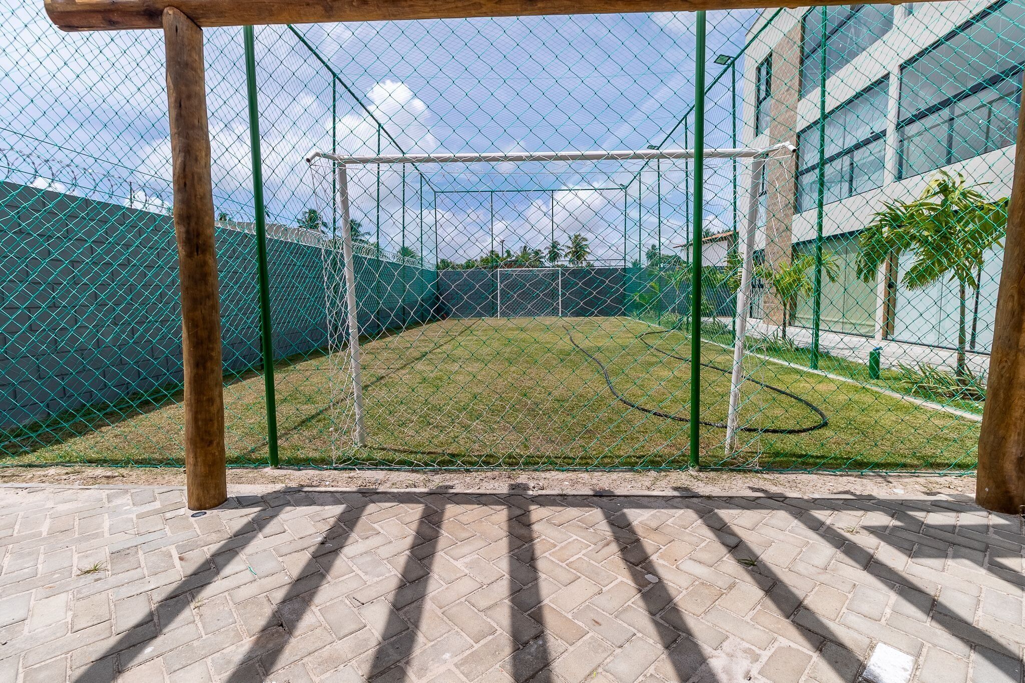 Um campo de críquete cercado por uma grade e uma rede de proteção, ladeado por uma área com piso de tijolos e um edifício com grandes janelas.