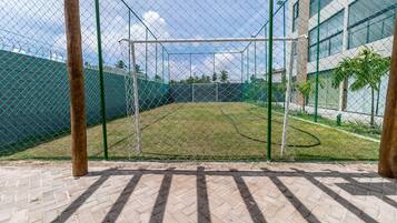 Um campo de críquete cercado por uma grade e uma rede de proteção, ladeado por uma área com piso de tijolos e um edifício com grandes janelas.