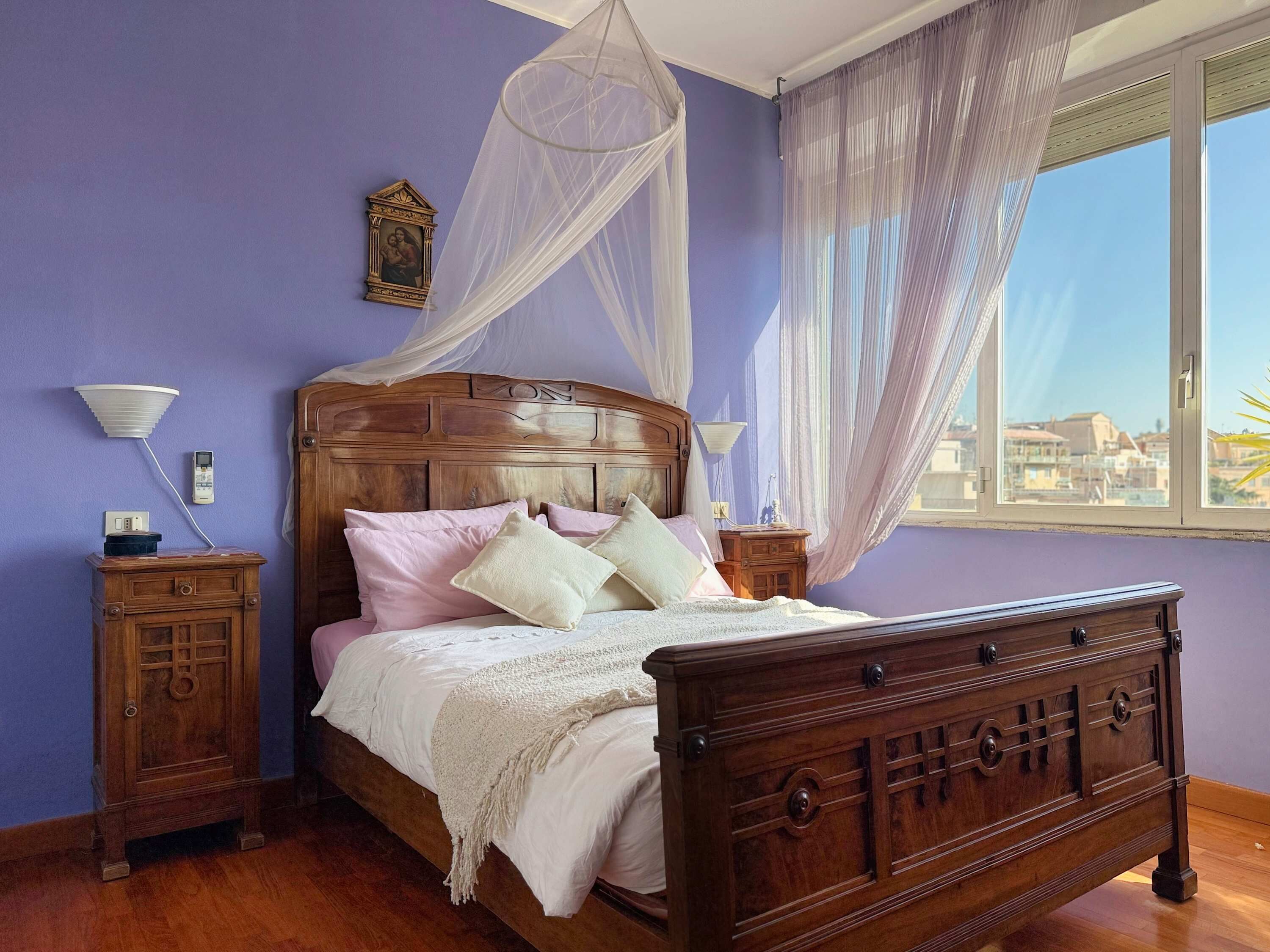 Une chambre avec un lit en bois, une table de chevet, une commode, une fenêtre avec des rideaux légers et une vue sur la ville.