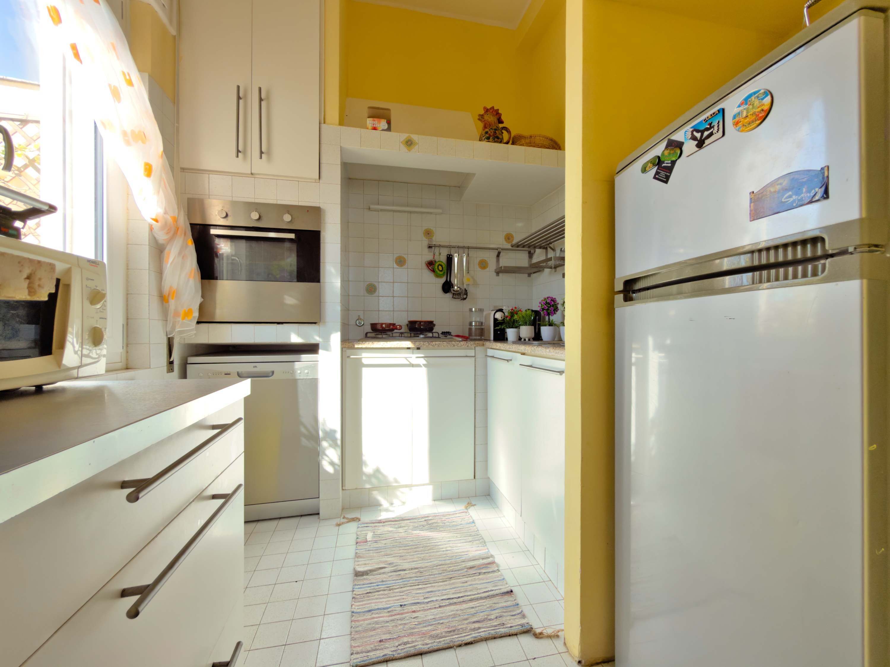 A kitchen with white cabinets, a microwave, an oven, and a refrigerator. There are potted plants on the counter and a rug on the floor.