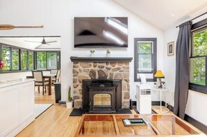 A modern living room with a stone fireplace, a flat-screen TV mounted on the wall, and a dining area visible through large windows. - Cozy Lake House Retreat. EV Charger, Modern Cabin. (Vernon Township)