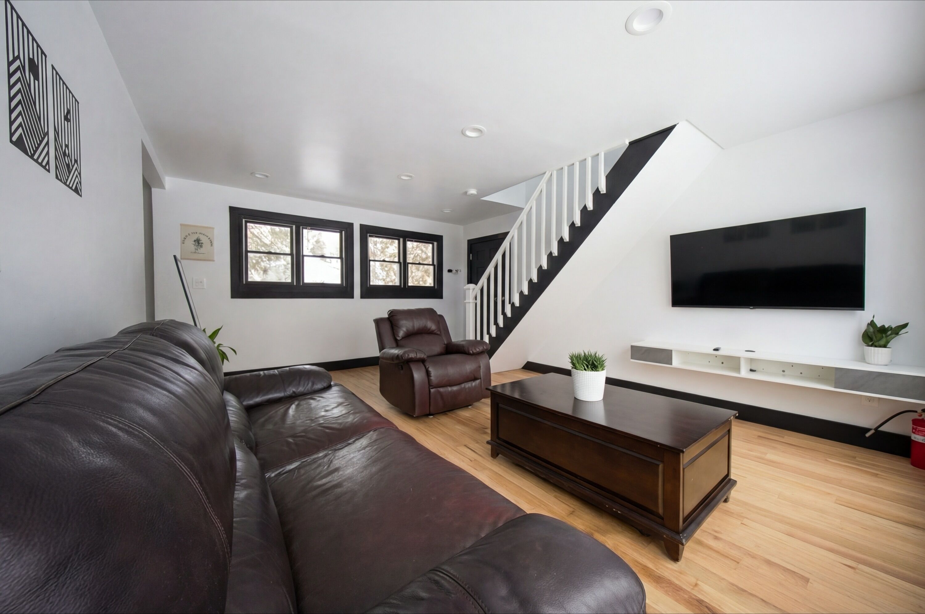 A modern living room with a leather sofa, a brown armchair, a wooden coffee table, a flat-screen TV mounted on the wall, and a staircase with black railings.