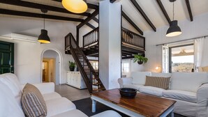 Amplia sala de estar con techo alto, vigas de madera, sofás blancos, una mesa de centro de madera y una escalera que lleva a un nivel superior.