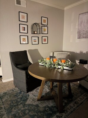 Uma mesa de madeira redonda com um arranjo floral, uma poltrona escura e um tapete estampado.