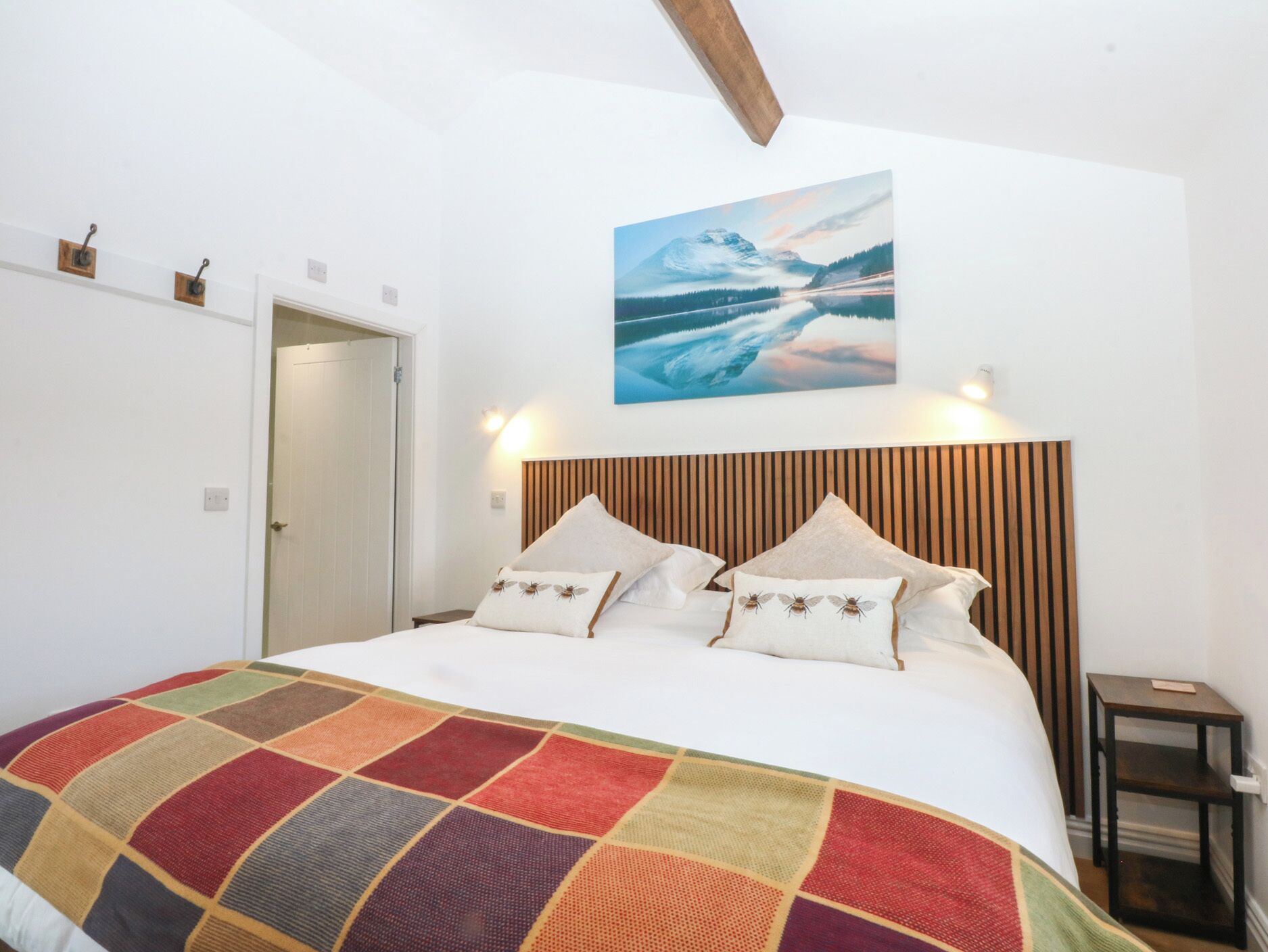 Une chambre à coucher avec un lit, une couverture colorée, une tête de lit en bois, un tableau représentant un paysage de montagne et une petite table près du lit.