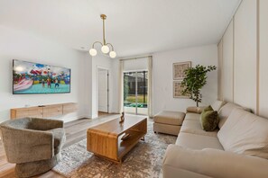 Ein modernes Wohnzimmer mit einem Flachbildfernseher, einem Holztisch, einem beigefarbenen Sofa und einem großen Fenster.