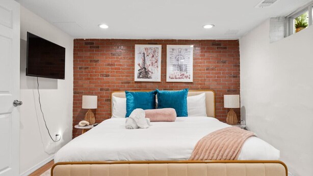 A bedroom with a brick wall, a bed with blue and pink pillows, a TV on the wall, and a bedside table with a lamp. - Family-Friendly 3BR Home Near DC (Arlington)