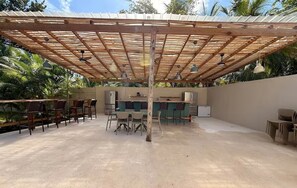 Un patio exterior con una pérgola de madera, mesas, sillas y una zona de bar.