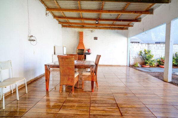 A covered patio area with a brick fireplace, a dining table with chairs, and a white chair. - Casa c Piscina Churrasqueira Perto do Rio Grande (Itapagipe)