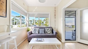 A room with a white table, a purple sofa with patterned cushions, a window with a view of trees, and a door with a metal gate. - Beauts - Pet Friendly - 5 Mins Walk to Beach (Culburra Beach)