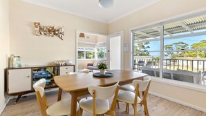 A dining area with a wooden table and chairs, a cabinet with items on top, and a view through large windows. - Beauts - Pet Friendly - 5 Mins Walk to Beach (Culburra Beach)