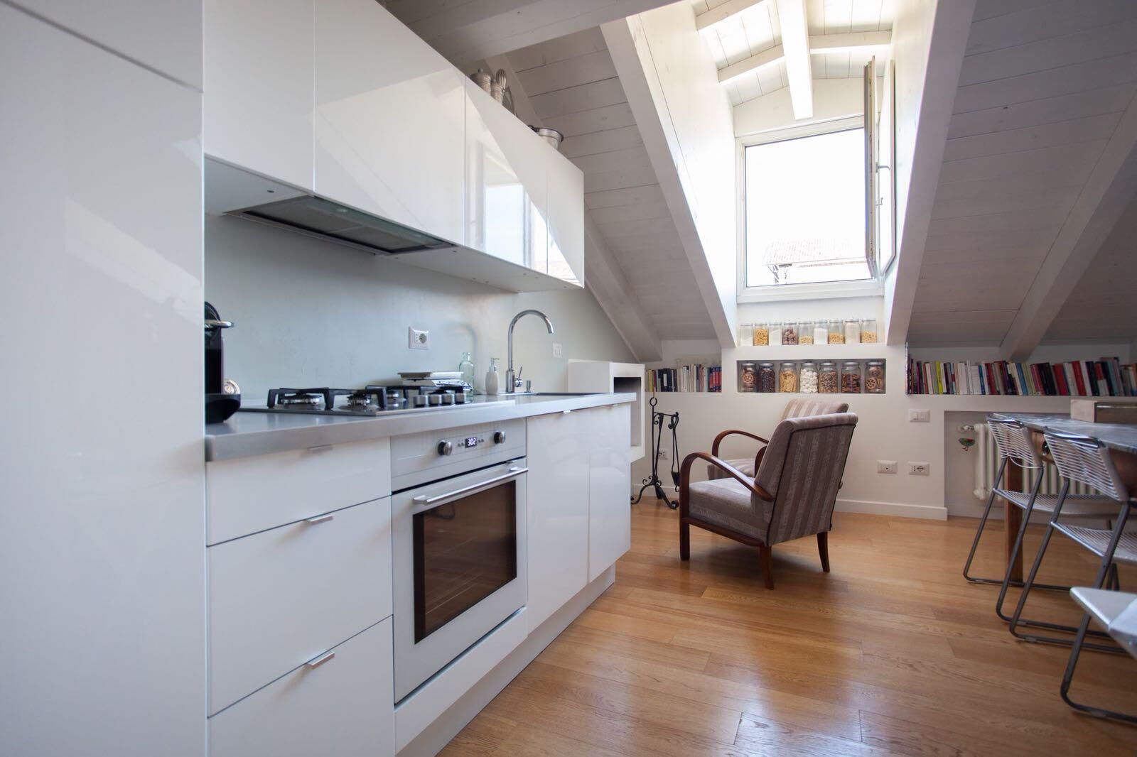 Een moderne keuken met witte kasten, een gasfornuis en een inbouwoven. Er is een eethoek met een stoel en een tafel, en een boekenkast vol boeken.