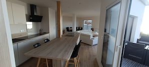 A modern kitchen with a wooden table and chairs, a dining area with a white sofa, and a large sliding glass door leading to an outdoor area.