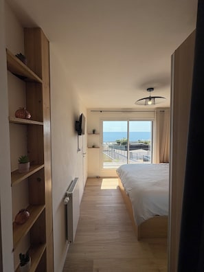 A bedroom with a bed, a radiator, wooden shelving, a television, and a view of the sea through a large window. - Villa Tregana Beach: comfort and immersion in an idyllic landscape (Locmaria-Plouzané)