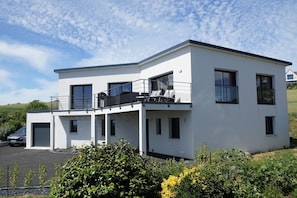 A modern two-story house with a balcony, a car parked in front, and lush greenery around.