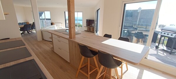 A modern kitchen with a wooden countertop, bar stools, and a view of the sea through large windows.