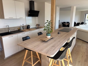 A modern kitchen with a central island, wooden stools, a potted plant, and a dining area with a sofa and chairs.